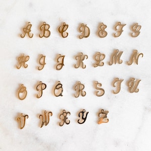 Pode incluir: Um conjunto de 26 pingentes de letras do alfabeto em metal dourado. Cada pingente é uma letra cursiva do alfabeto, de A a Z.