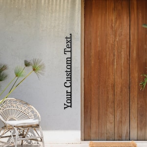 Könnte beinhalten: Eine Holztür mit einer braunen Fußmatte davor. An der weißen Wand steht der Text "Your Custom Text". Rechts und links von der Tür stehen zwei Topfpflanzen.