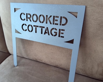 May include: A gray metal sign with the words "CROOKED COTTAGE" cut out. The sign is attached to a metal stake.