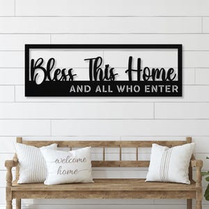 Pode incluir: Uma placa de metal preta com as palavras "Bless This Home" e "And All Who Enter" em uma fonte decorativa. A placa é montada em uma parede branca de ripas acima de um banco de madeira com duas almofadas decorativas.