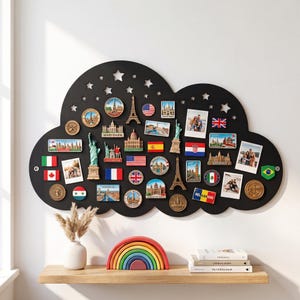 Può includere: Una lavagna magnetica nera a forma di nuvola con ritagli a forma di stella nella parte superiore. La lavagna è ricoperta da vari magneti con bandiere internazionali, souvenir di monumenti e diverse fotografie rettangolari attaccate.