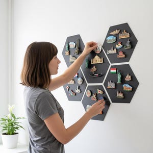 Puede incluir: Una persona organizando imanes de recuerdo de viaje en una pizarra magnética hexagonal negra. Los imanes representan monumentos famosos como la Torre Eiffel, la Estatua de la Libertad y la Torre de Pisa. La pizarra está montada en una pared blanca.
