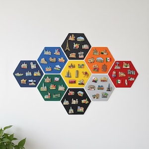 Puede incluir: Una colección de imanes hexagonales coloridos, cada uno con miniaturas de monumentos e iconos de varios países. Los imanes están dispuestos en una pared blanca, mostrando una exhibición con temática de viajes. Incluye la Torre Eiffel, el Big Ben y el Coliseo.