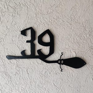 Könnte beinhalten: Schwarzes Hausnummernschild aus Metall mit der Nummer 39 in einer stilisierten Schriftart. Die Nummer ist an einem schwarzen Besen aus Metall befestigt.