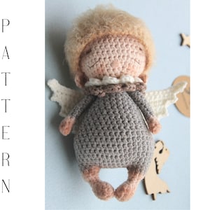 Può includere: Una bambola angelo in crochet con un vestito grigio, ali bianche e un viso marrone chiaro. La bambola ha capelli ricci castano chiaro e un colletto in crochet bianco.