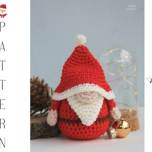 May include: A crocheted Santa Claus gnome with a red hat, white beard, and brown belt. The gnome is sitting on a wooden surface with a gold bauble nearby. The text "Ho ho ho!" is in the background.