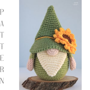 May include: A green crocheted gnome with a white beard and a yellow sunflower on its hat. The gnome is sitting on a wooden slice.