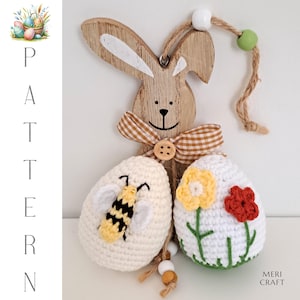 May include: A wooden bunny cutout with a brown and white checkered bow, holding two crocheted Easter eggs. One egg has a bee design and the other has a flower design.