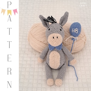 Puede incluir: Un juguete de burro de crochet gris con una pajarita azul que sostiene un globo azul con las letras "HB" en él. El burro está sentado sobre una superficie blanca con una bola de lana en el fondo.
