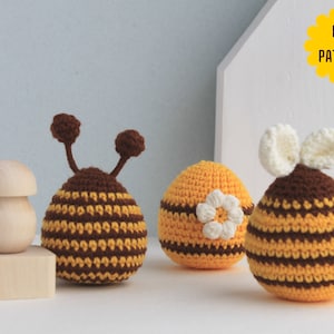 May include: Three crocheted bee toys, one with brown and yellow stripes, one with brown and yellow stripes and a white flower, and one with brown and yellow stripes and white wings. The toys are on a white surface with a wooden block and a wooden mushroom.