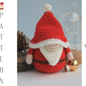 May include: Crocheted Santa Claus gnome ornament with a red hat, white beard, and brown belt. The gnome is sitting on a wooden surface with a gold ornament in the background. The text "Ho ho ho!" is written in green on a white background.
