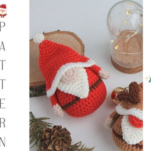 May include: Two crocheted Christmas decorations, one a Santa Claus with a red hat and white beard, and the other a reindeer with brown antlers and a red nose. The decorations are sitting on a wooden surface with a pine cone and a glass jar with lights in the background. The text "Ho ho ho!" is written in green on a white background.