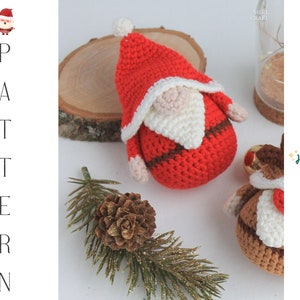 May include: A crocheted Santa Claus ornament with a red hat, white beard, and brown belt. The ornament is sitting on a wooden surface with a pine branch and a pine cone.