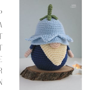 May include: A blue and white crocheted gnome wearing a blue flower hat with a green stem. The gnome is sitting on a wooden slice.