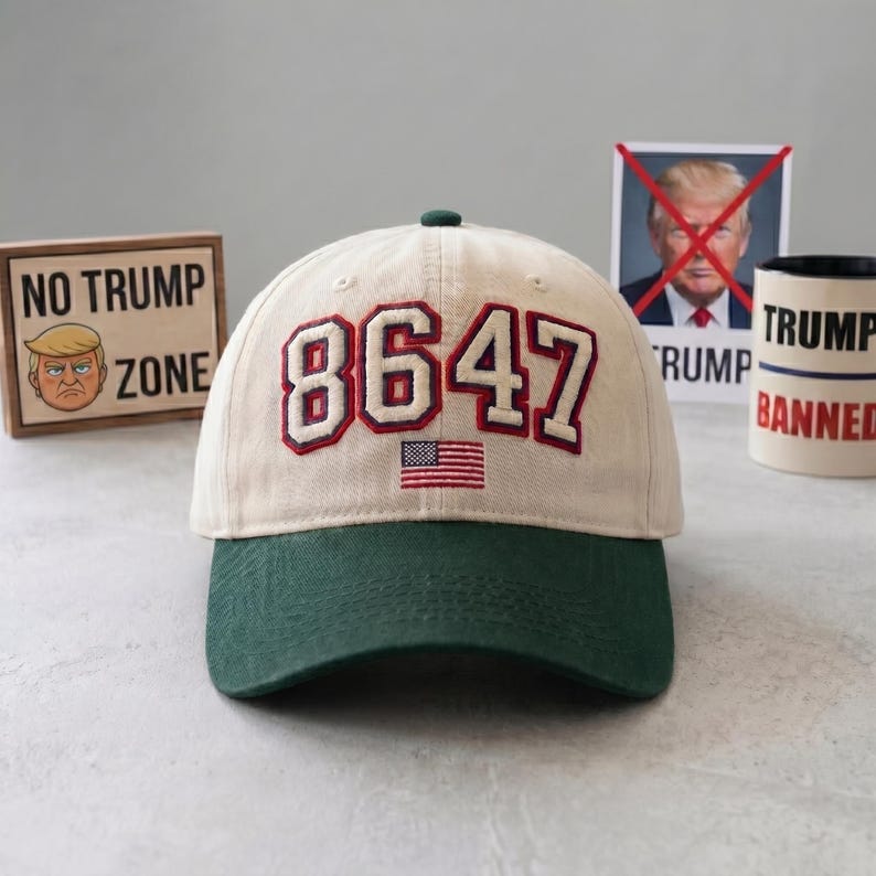 May include: A beige and green baseball cap with the number "8647" embroidered in red and white, along with a small American flag. The cap has a green brim and a green button on top. In the background are anti-Trump signs.