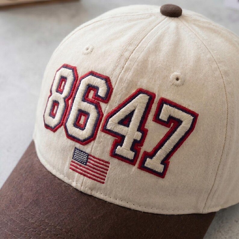 May include: A beige baseball cap with a brown brim and top button. The cap features the number "8647" embroidered in white with red and blue accents, along with a small American flag.