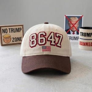 May include: A beige baseball cap with a brown brim, embroidered with the number "8647" in red and white, and a small American flag. The cap is part of a collection of anti-Trump merchandise.