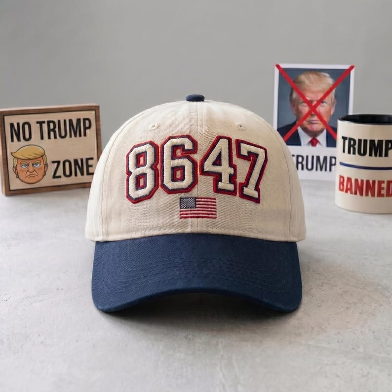 May include: A beige and navy blue baseball cap with the number "8647" embroidered in red and white, along with a small American flag. The cap is part of a collection of anti-Trump merchandise.