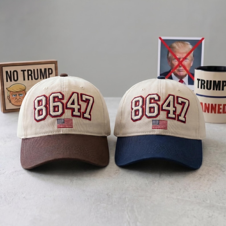 May include: Two baseball caps with the number "8647" embroidered in red and white, along with a small American flag. One cap has a brown brim, the other a navy blue brim. The caps are on a table with anti-Trump imagery.