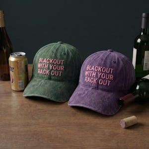 May include: Two baseball caps, one green and one purple, with the text "BLACKOUT WITH YOUR RACK OUT" embroidered in pink. The caps are on a wooden surface, with bottles and a can of beer in the background.