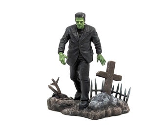 May include: A green-skinned Frankenstein figure in a black suit stands in a graveyard setting. The figure is walking towards a wooden cross with a metal fence around it. The figure is part of a collectible action figure set.