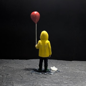 Puede incluir: Figura de una persona con impermeable amarillo con capucha sosteniendo un globo rojo. La figura está sobre una superficie oscura y texturizada con un barco de papel cerca, sobre un fondo negro.