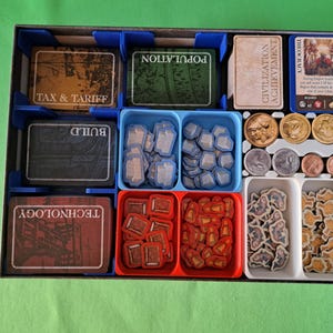 Op de afbeelding: Een bordspel organizer met diverse spelonderdelen. De organizer heeft blauwe, rode en witte bakjes met kaarten, fiches en munten. Kaarten zijn gelabeld met "TAX & TARIFF", "POPULATION", "BUILD", "TECHNOLOGY", "CIVILIZATION ACHIEVEMENT" en "THEOCRACY".