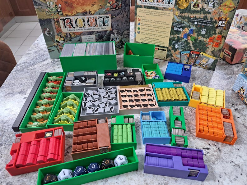 Op de afbeelding: Het bordspel "Root" met diverse spelonderdelen georganiseerd in gekleurde bakjes. De speldoos is zichtbaar, samen met kaarten en dobbelstenen. De spelstukken zijn in tinten groen, rood, oranje, geel, blauw en paars.