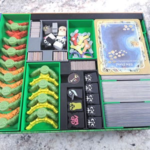 Op de afbeelding: Een groene en blauwe bordspelorganizer met diverse spelonderdelen. De organizer heeft verschillende compartimenten gevuld met kleurrijke spelcomponenten, waaronder kaarten, fiches en karakterfiguren. De spelstukken zijn in tinten rood, oranje, geel en zwart.