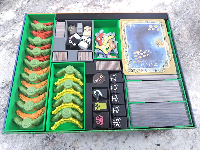 Op de afbeelding: Een groene en blauwe bordspelorganizer met diverse spelonderdelen. De organizer heeft verschillende compartimenten gevuld met kleurrijke spelcomponenten, waaronder kaarten, fiches en karakterfiguren. De spelstukken zijn in tinten rood, oranje, geel en zwart.