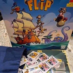 Könnte beinhalten: Ein Brettspiel namens "Captain Flip" mit einem blauen, grünen und weißen Spielbrett und einem Stapel bunter Karten. Die Spielschachtel zeigt eine Comic-Illustration eines Schiffes mit einem Piratenkapitän und einem Monsterkraken.