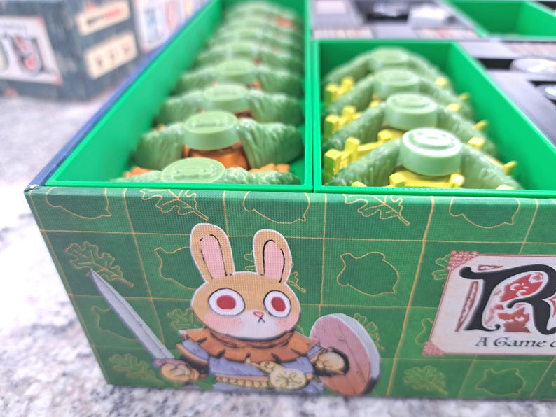 Op de afbeelding: Bordspeldoos met een groen en blauw ontwerp. De doos heeft een cartoon konijn met een zwaard en schild. Binnenin zitten groene en gele spelstukken. De speltitel "Root" is zichtbaar op de doos.