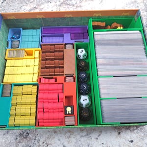 Op de afbeelding: Een bordspel organizer met meerdere compartimenten in blauw, geel, rood en paars. De organizer bevat spelstukken, kaarten en dobbelstenen. De groene doos heeft een patroon aan de binnenkant.