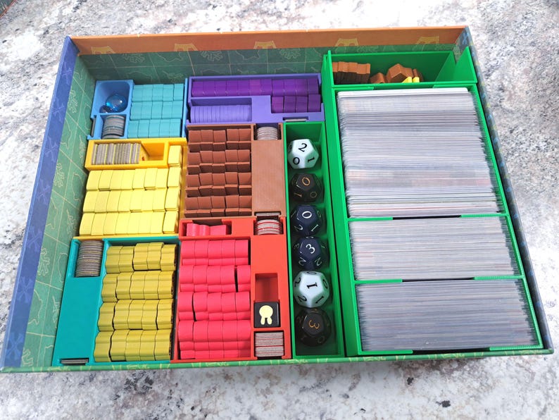 Op de afbeelding: Een bordspel organizer met meerdere compartimenten in blauw, geel, rood en paars. De organizer bevat spelstukken, kaarten en dobbelstenen. De groene doos heeft een patroon aan de binnenkant.