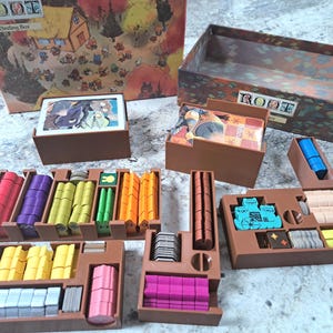 Peut inclure: Un ensemble d'organisateur de jeu de société pour le jeu "Root". L'ensemble comprend des boîtes et des plateaux de rangement marron remplis de pièces de jeu, de cartes et de jetons colorés. La boîte de jeu est visible en arrière-plan, avec le titre du jeu "Root" imprimé dessus.