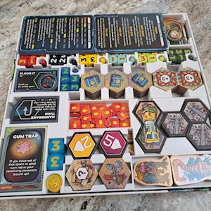 May include: A white plastic game organiser with compartments for storing game pieces. The organiser contains dice, cards, tokens, and tiles. The game pieces are themed around a science fiction or fantasy setting. The text on the cards reads "Floor It", "Turn", "Handbrake", and "Gum Trap".