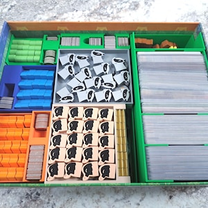 Op de afbeelding: Een bordspel organizer met diverse compartimenten gevuld met spelstukken. De compartimenten zijn in verschillende kleuren, waaronder groen, blauw, oranje en grijs. De spelstukken omvatten kaarten, houten fiches en kleine beeldjes.