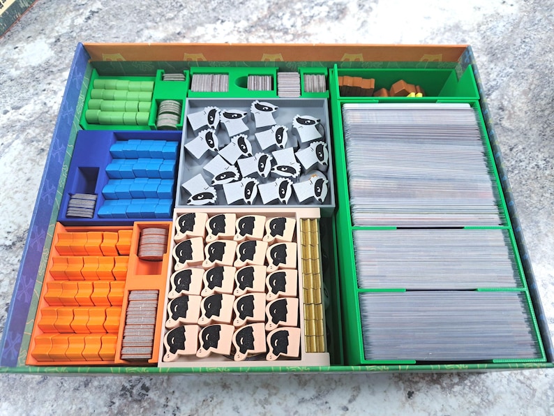 Op de afbeelding: Een bordspel organizer met diverse compartimenten gevuld met spelstukken. De compartimenten zijn in verschillende kleuren, waaronder groen, blauw, oranje en grijs. De spelstukken omvatten kaarten, houten fiches en kleine beeldjes.