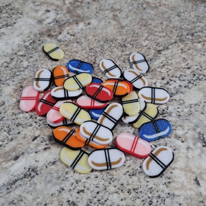 Op de afbeelding: Een stapel kleurrijke plastic pillen-vormige bedels in verschillende kleuren, waaronder rood, oranje, geel, blauw en wit. De bedels hebben zwarte lijnen en witte accenten.