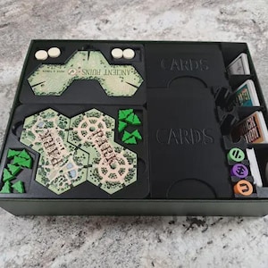 May include: A black board game organiser insert containing hexagonal park tiles, small green tree tokens, white circular tokens, and stacks of cards labelled CARDS. The insert features dedicated compartments for game components and organised storage.