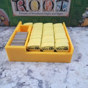 Op de afbeelding: Een gele plastic spelorganizer met stapels gele houten tokens met zwart bedrukte gezichten en een stapel kleine grijze kaarten. De speldoos op de achtergrond leest "Root: A Game of Woodland Might and Right."