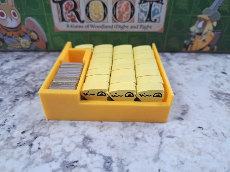 Op de afbeelding: Een gele plastic spelorganizer met stapels gele houten tokens met zwart bedrukte gezichten en een stapel kleine grijze kaarten. De speldoos op de achtergrond leest "Root: A Game of Woodland Might and Right."