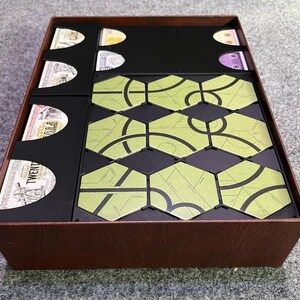 May include: A brown game box with black compartments holding game components. The box contains green hexagonal tiles with black designs and various game cards. The box is designed to organize and store board game pieces.