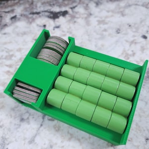 Op de afbeelding: Een felgroene 3D-geprinte spelstukkenorganizer. De organizer heeft compartimenten voor ronde, platte spelstukken en cilindrische spelstukken. De tokens zijn grijs en de cilindrische stukken lichtgroen. De organizer staat op een gespikkelde ondergrond.