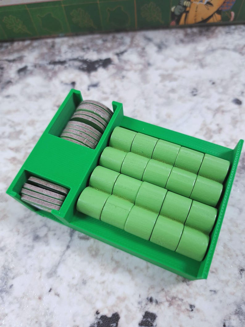 Op de afbeelding: Een felgroene 3D-geprinte spelstukkenorganizer. De organizer heeft compartimenten voor ronde, platte spelstukken en cilindrische spelstukken. De tokens zijn grijs en de cilindrische stukken lichtgroen. De organizer staat op een gespikkelde ondergrond.