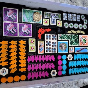 Puede incluir: Un organizador de juegos de mesa lleno de varios componentes del juego. Incluye piezas de jugador naranjas, rosas y azules, cartas y fichas. Los componentes están dispuestos en bandejas negras dentro de una caja blanca.