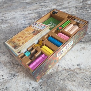 May include: A Root board game insert with multiple compartments for organizing game pieces. The insert is made of brown cardboard and has a printed design of leaves and branches. The insert is filled with various game pieces, including wooden tokens, cards, and dice.