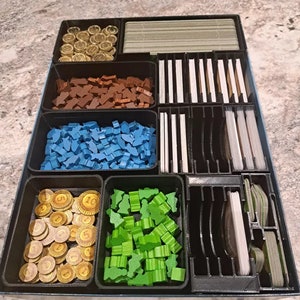 May include: A black organizer with multiple compartments for storing game pieces and cards. The compartments contain various colored wooden pieces, gold coins, and cards.