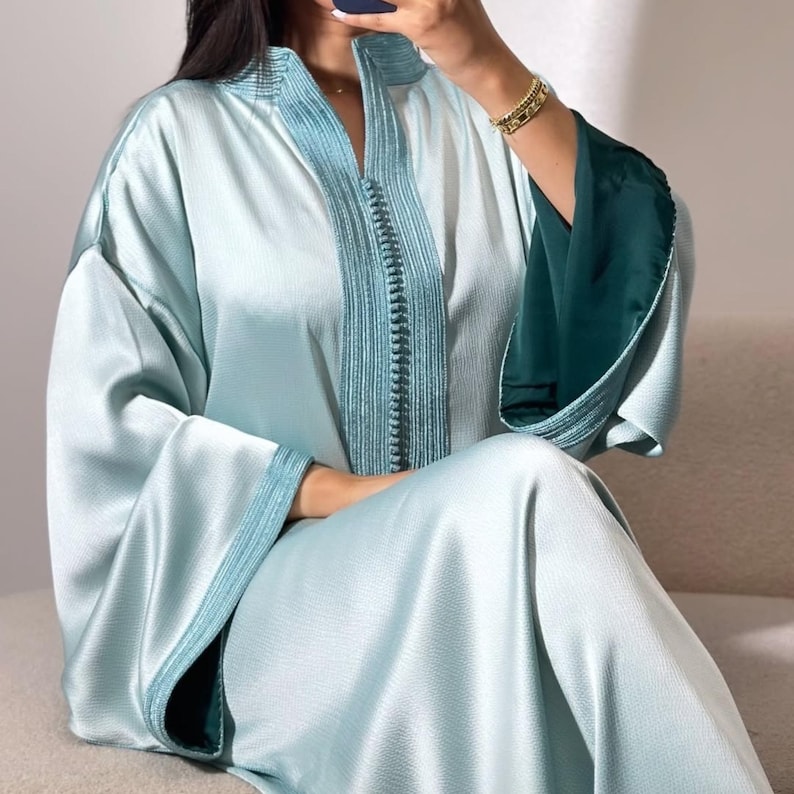 May include: A light blue satin robe with teal trim and a matching teal embroidered design down the front. The sleeves have a dark teal lining. The robe has a high collar and is worn by someone sitting down.