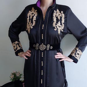 Puede incluir: Un vestido de sat&eacute;n negro con bordados dorados y un cintur&oacute;n de terciopelo negro. El vestido tiene un cuello alto y mangas largas. El bordado es intrincado y presenta dise&ntilde;os florales.